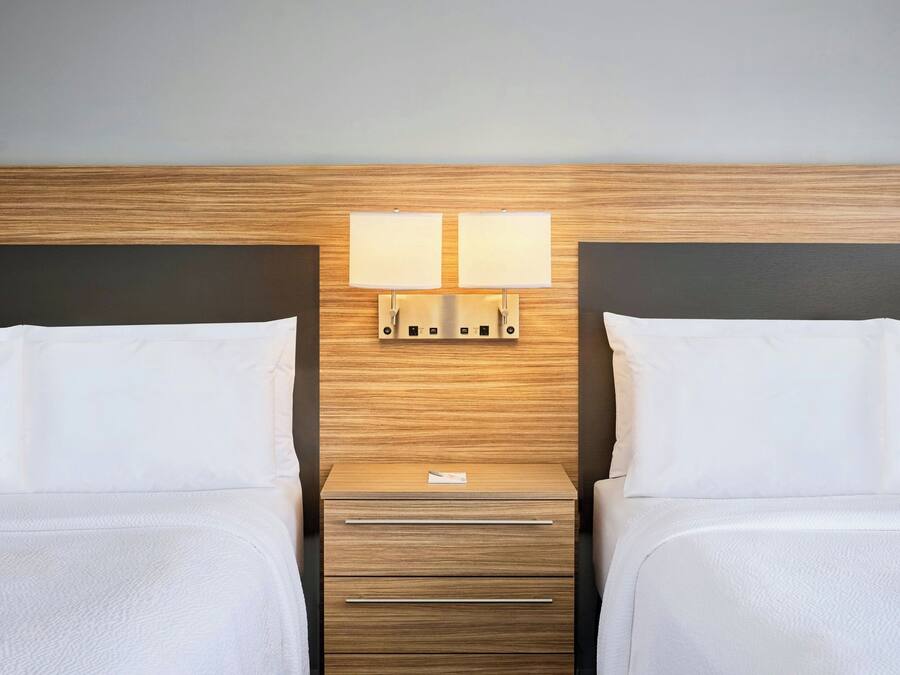 A hotel room with two beds, a wooden headboard, a nightstand with a lamp, and a drawer unit.