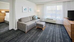 Een hotelkamer met een grijze bank, een houten kast, een flatscreen televisie en een groot raam met gordijnen.