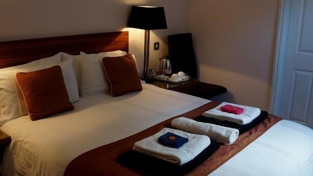 Een keurig opgemaakt bed met witte lakens en bruine kussens, een nachtkastje met een lamp en een dienblad met opgevouwen handdoeken.