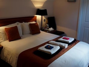 Una cama bien hecha con sábanas blancas y almohadas marrones, una mesita de noche con una lámpara, y una bandeja con toallas dobladas.