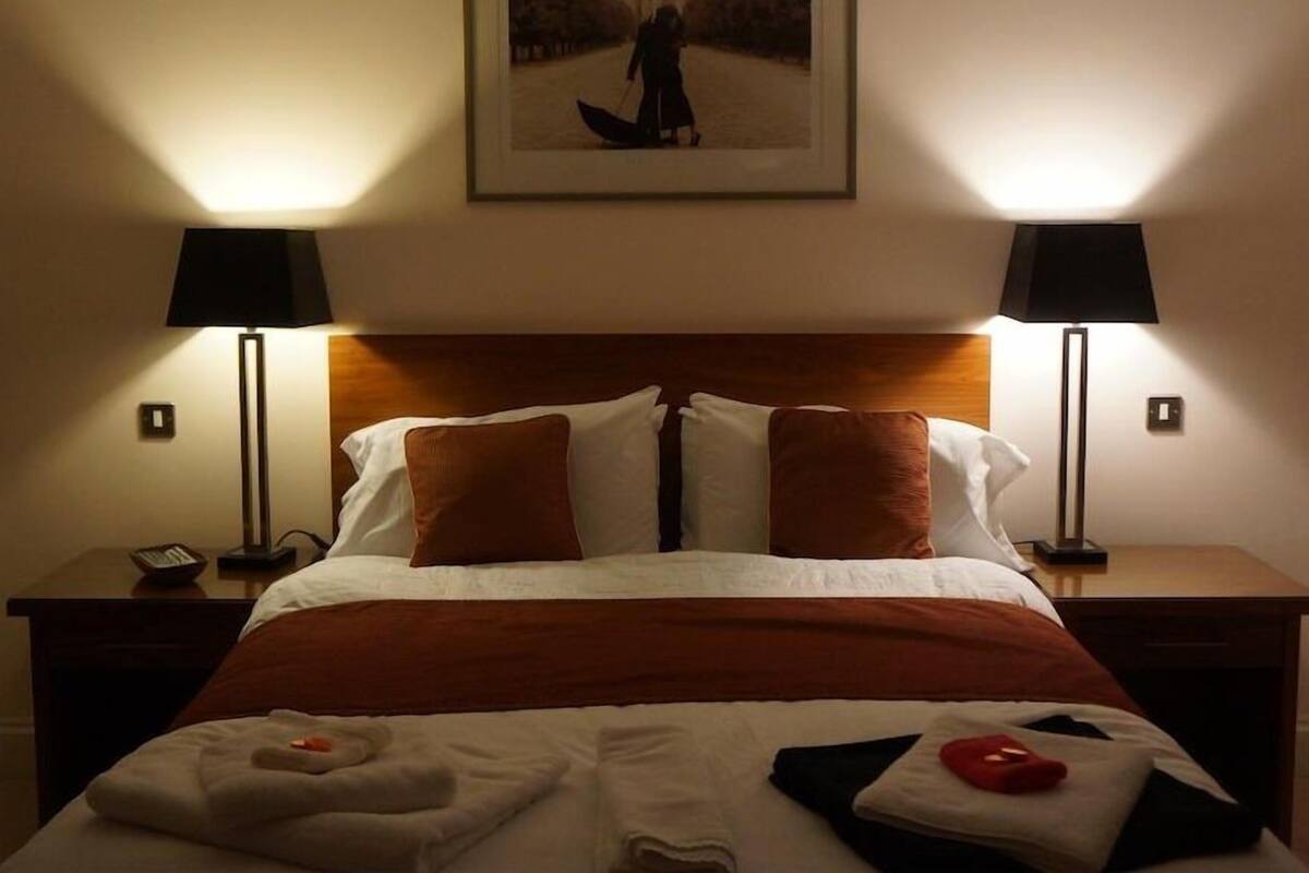A hotel room with a large bed, two bedside tables, a nightstand, a framed picture on the wall, and two floor lamps.