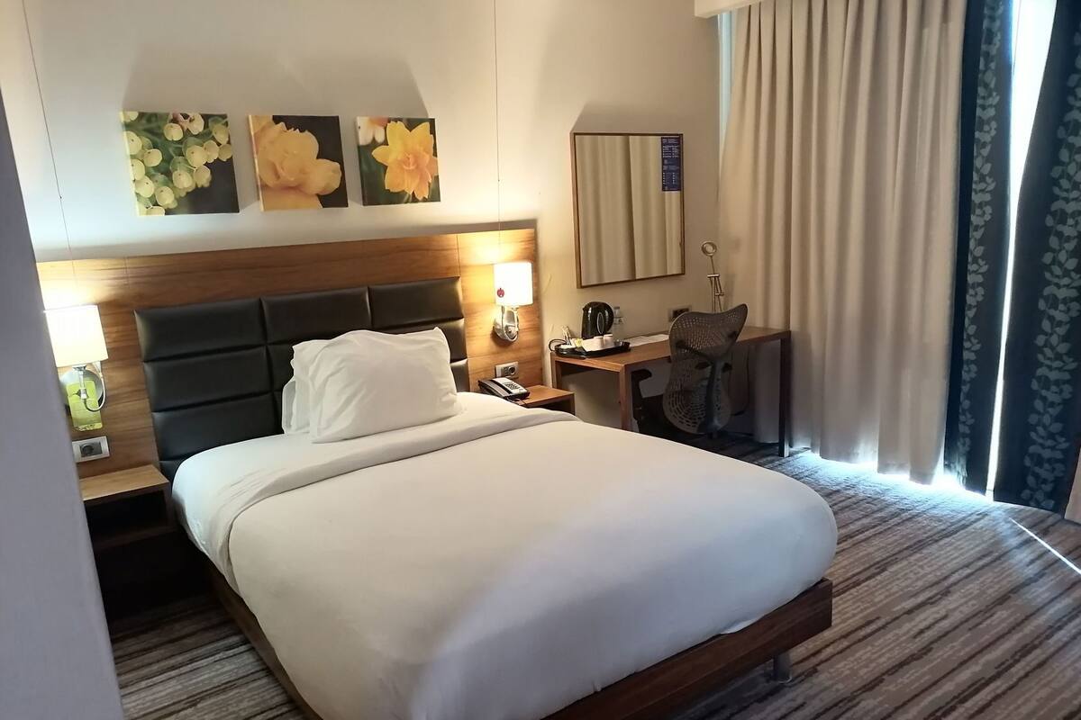 A hotel room with a large bed, a desk with a chair, a nightstand, a lamp, and a window with curtains.