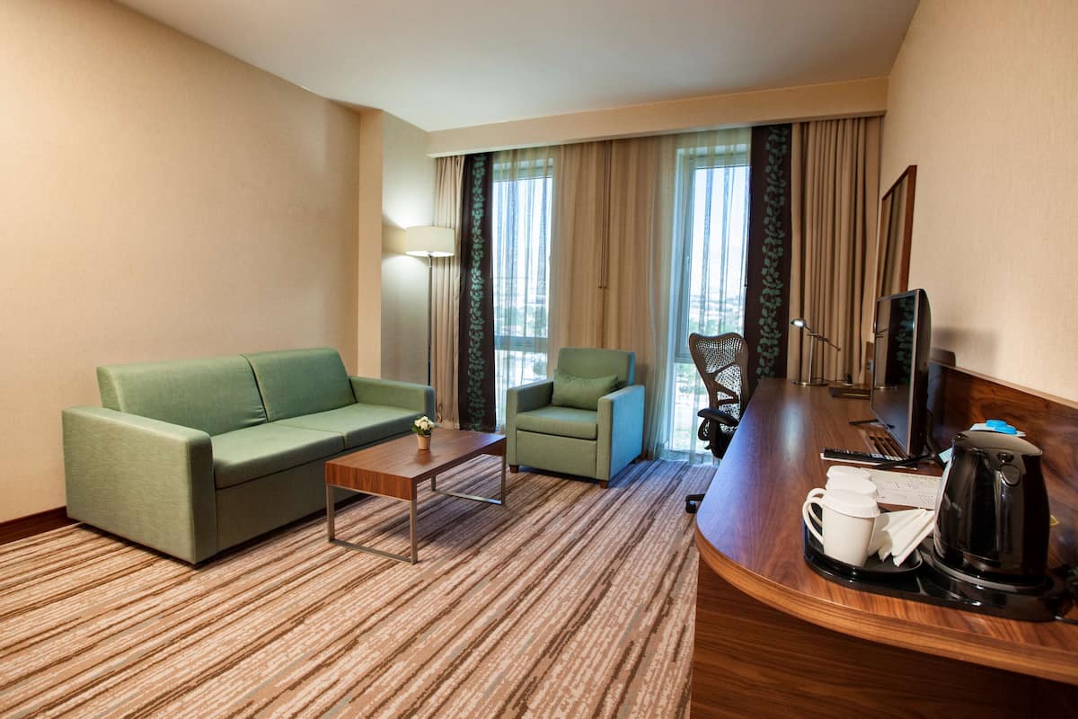 A hotel room with a green sofa, a small armchair, a wooden coffee table, a desk with a computer, a coffee maker, and a TV.
