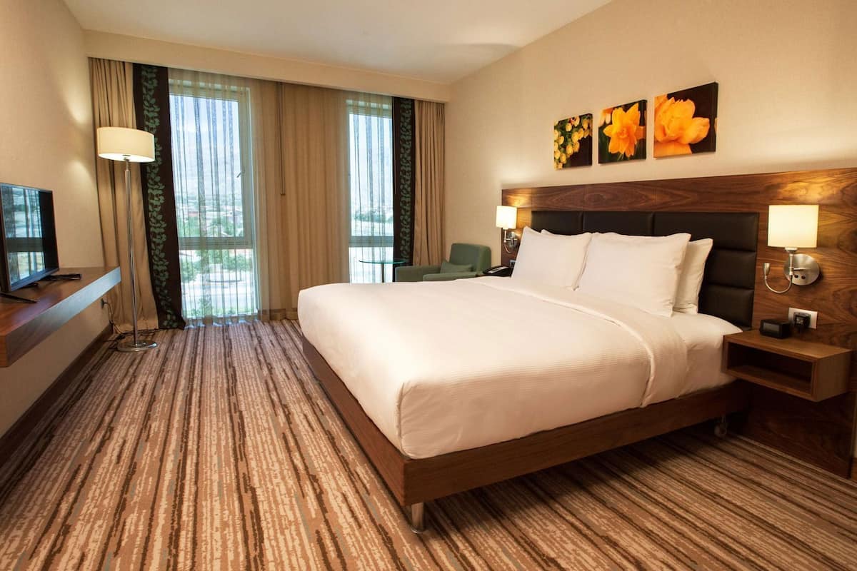 A hotel room with a bed, a desk with a TV, a chair, and a window with curtains.