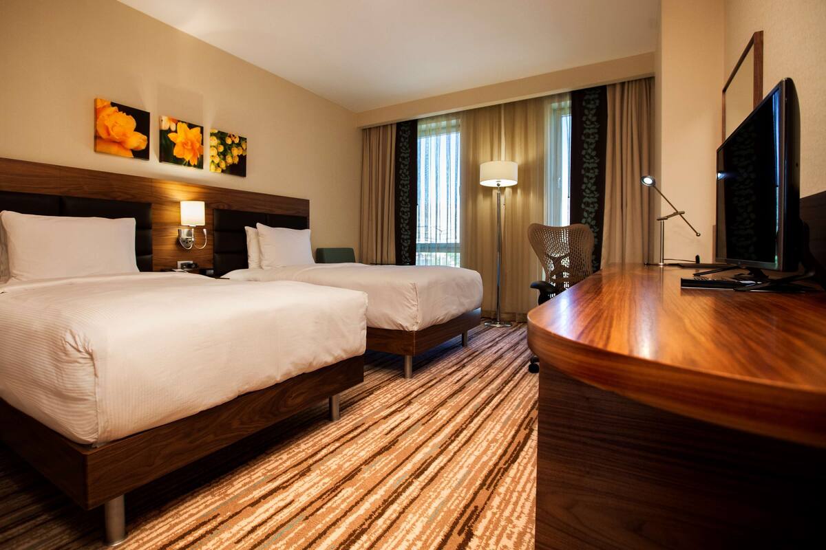 A hotel room with two beds, a desk, a TV, and a window with curtains.