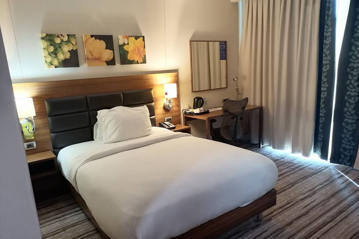 A hotel room with a large bed, a desk, a chair, a lamp, and artwork on the wall.