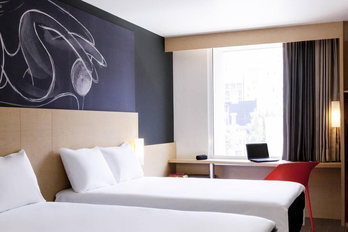 Habitación de hotel con dos camas, un ordenador portátil sobre una mesita, una silla roja y un cuadro abstracto grande en la pared.