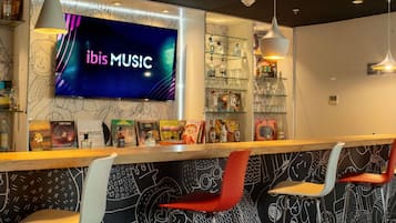 Zona de bar con televisor de pantalla plana mostrando 'ibis MUSIC' y varias botellas y vasos en estantes.