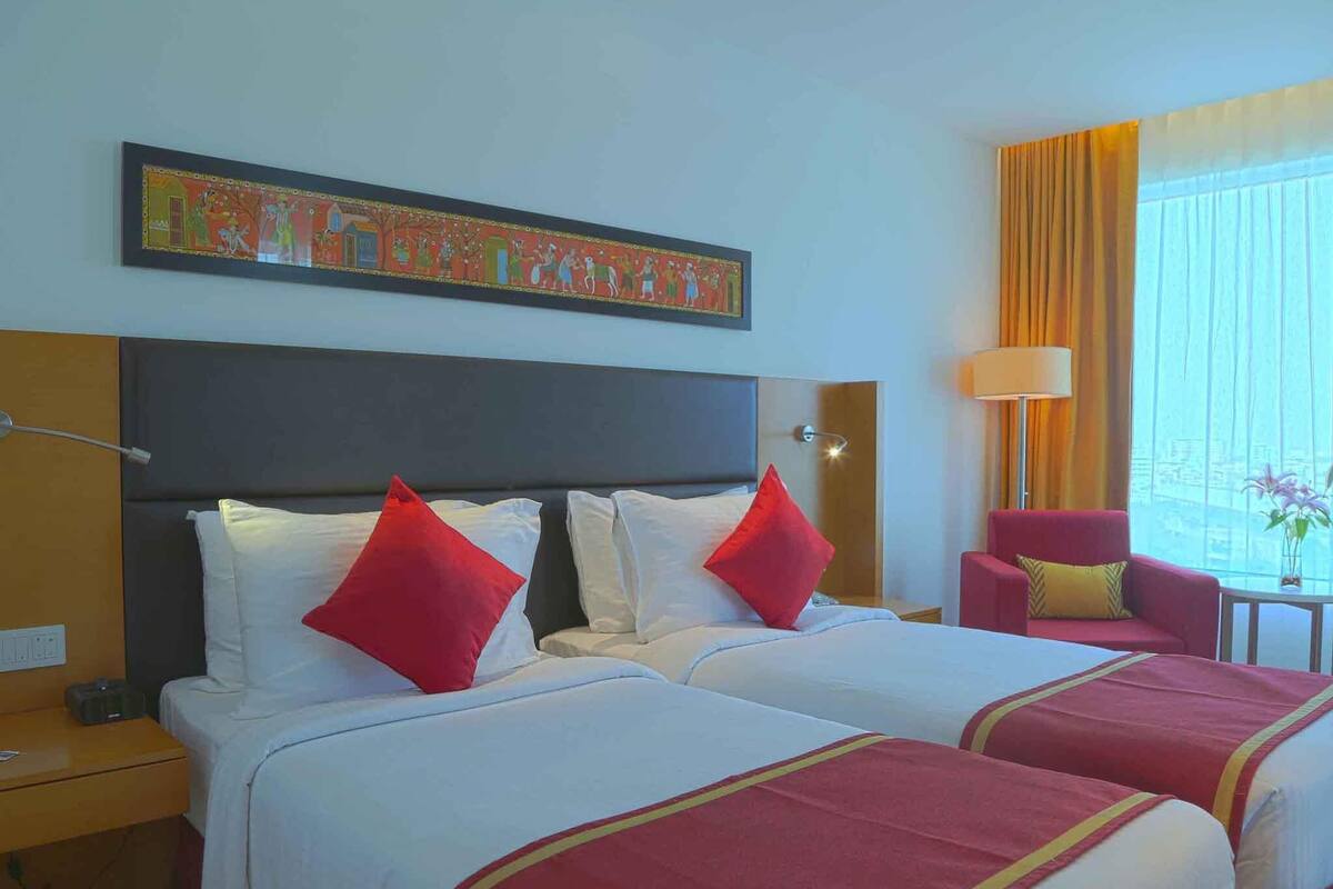 Une chambre d’hôtel avec deux lits, une décoration aux couleurs rouge et blanche, un grand tableau au-dessus des lits, une fenêtre avec des rideaux et une petite table avec une lampe.