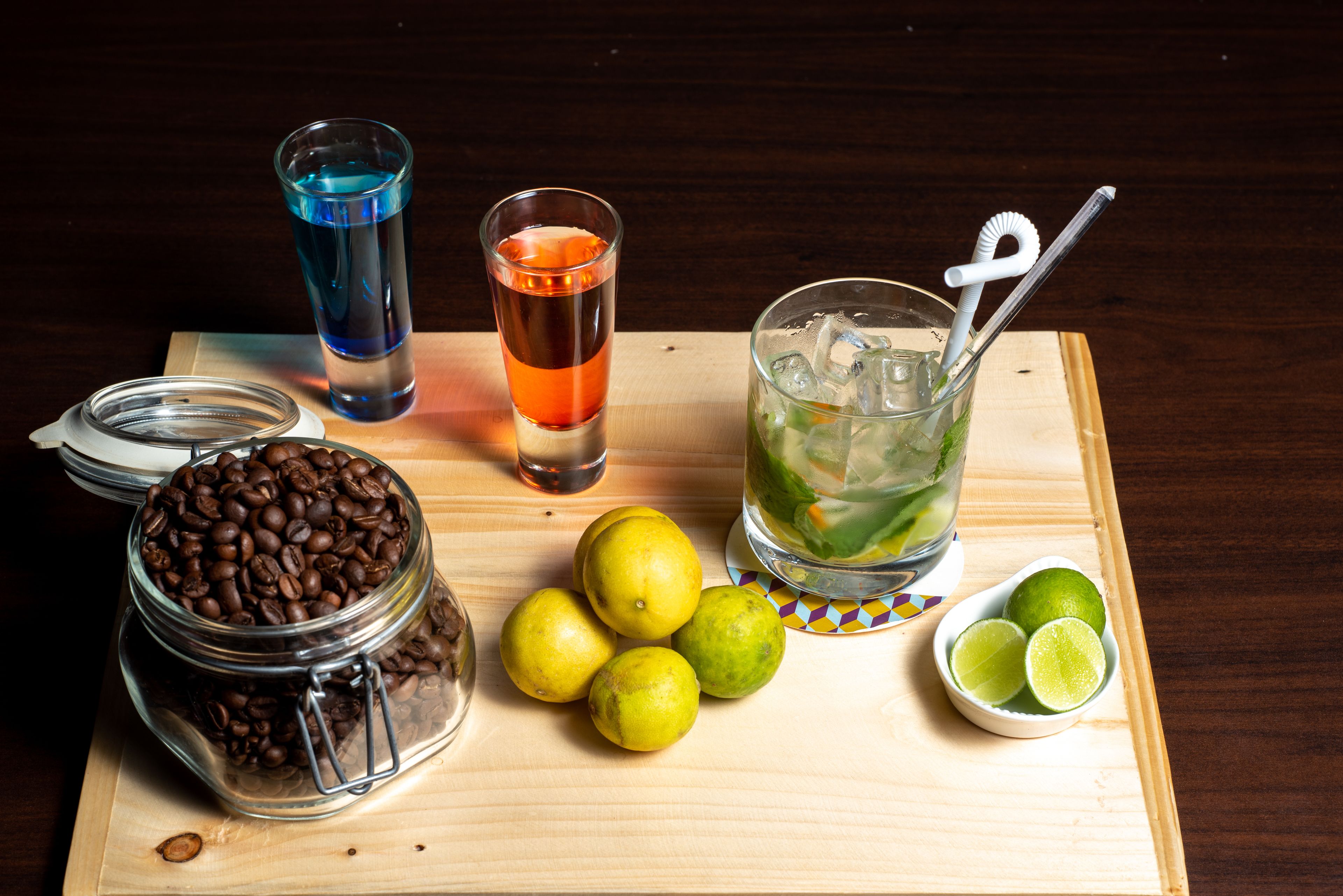 A wooden table with a glass jar of coffee beans, two glasses of beverages, and several limes.