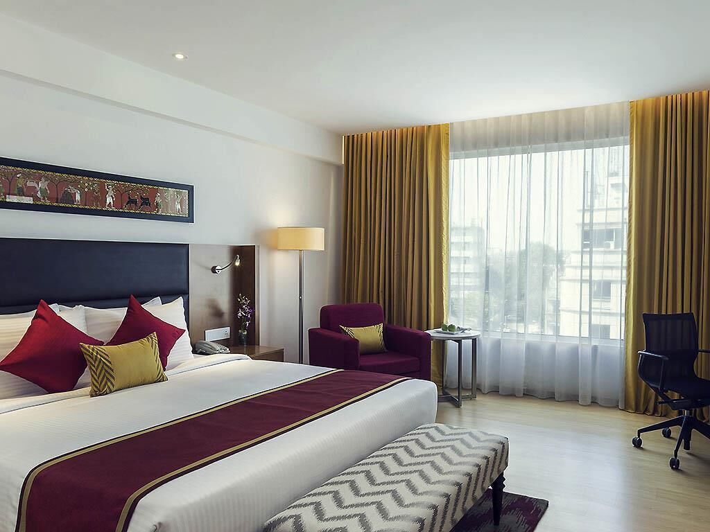 A hotel room with a large bed, a desk, a chair, and a view of the city through large windows.