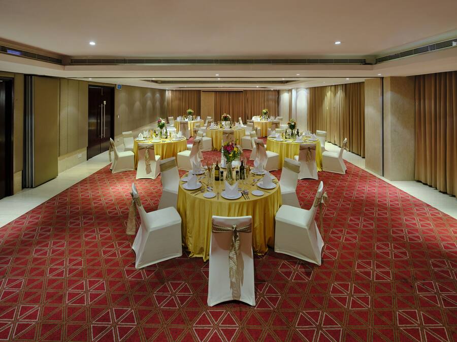 A banquet hall with round tables set for dining, white chairs, and a red patterned carpet.
