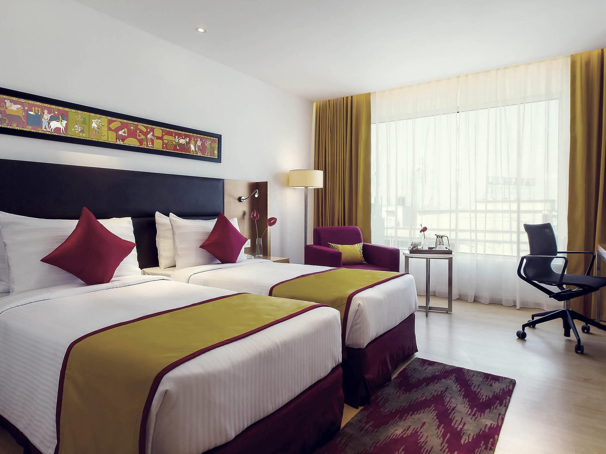Ein Hotelzimmer mit zwei Betten, einem Sessel, einem kleinen Tisch und einem großen Fenster mit Vorhängen.