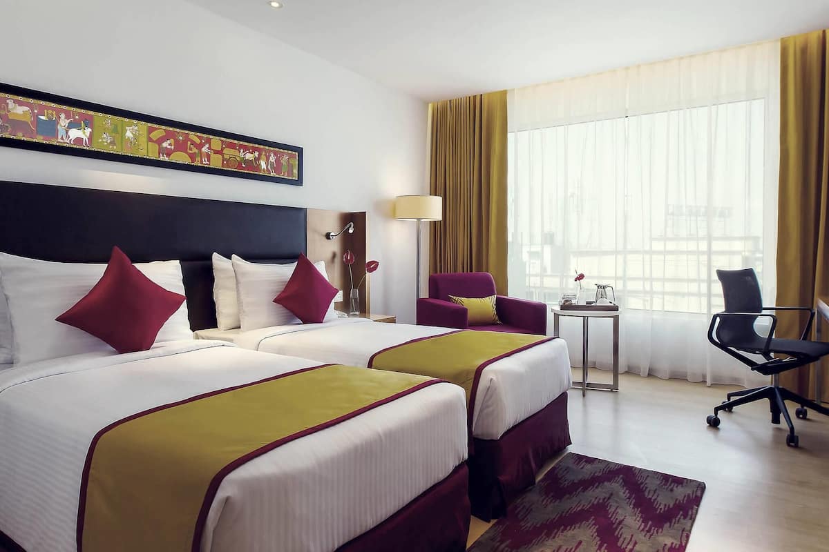 Ein Hotelzimmer mit zwei Betten, einem Sessel, einem kleinen Tisch und einem großen Fenster mit Vorhängen.