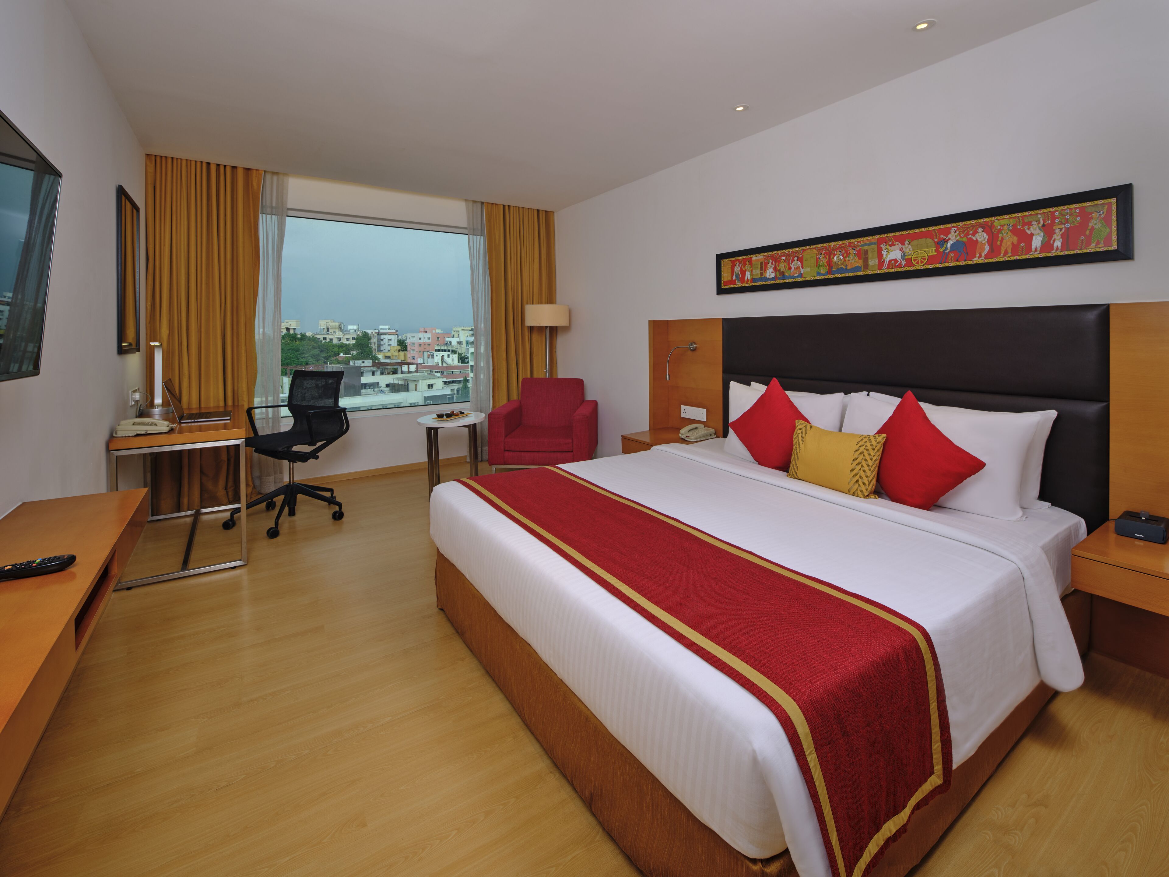 A hotel room with a large bed, a desk with a chair, a red armchair, a lamp, and a painting on the wall.