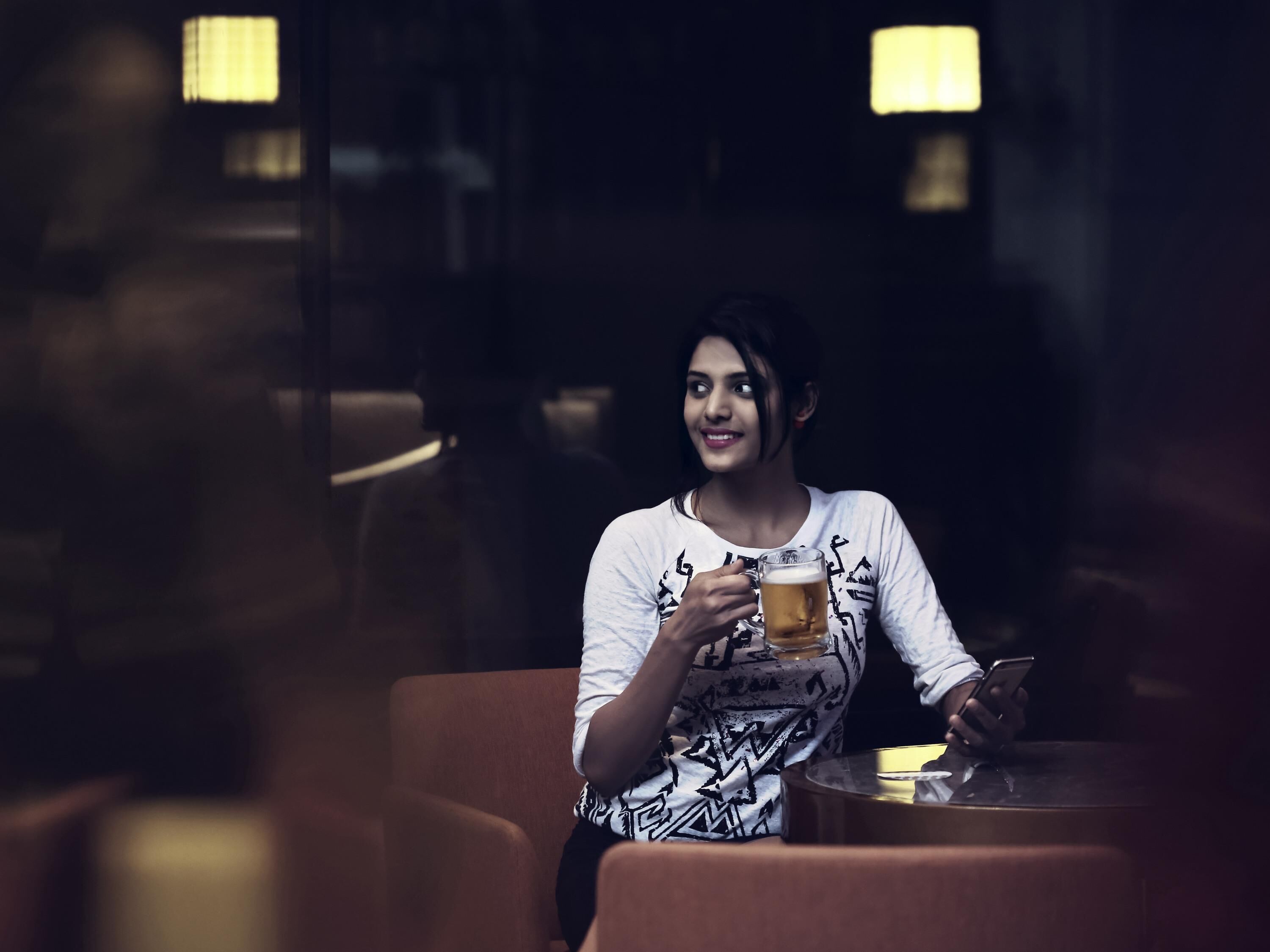 Una persona sentada a una mesa con una cerveza, sosteniendo un teléfono inteligente.