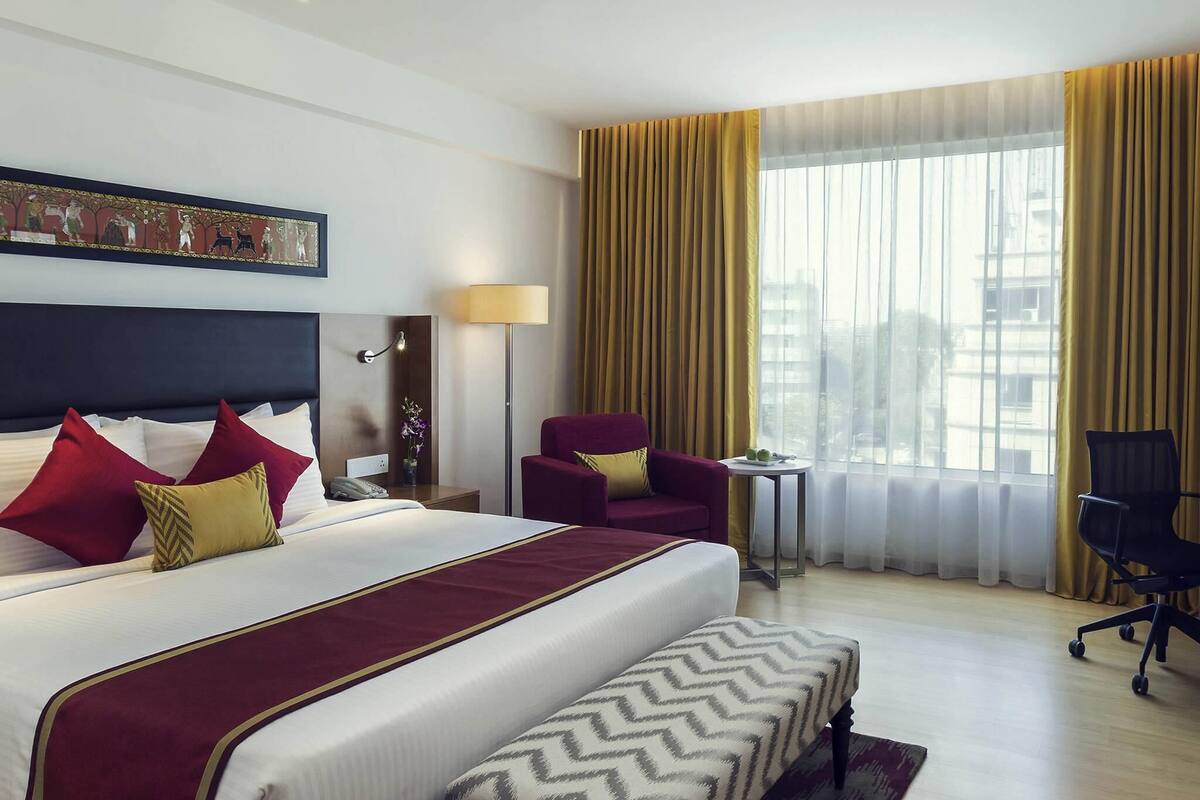 Une chambre d’hôtel avec un grand lit, un bureau, deux chaises, une lampe et une fenêtre avec des rideaux.