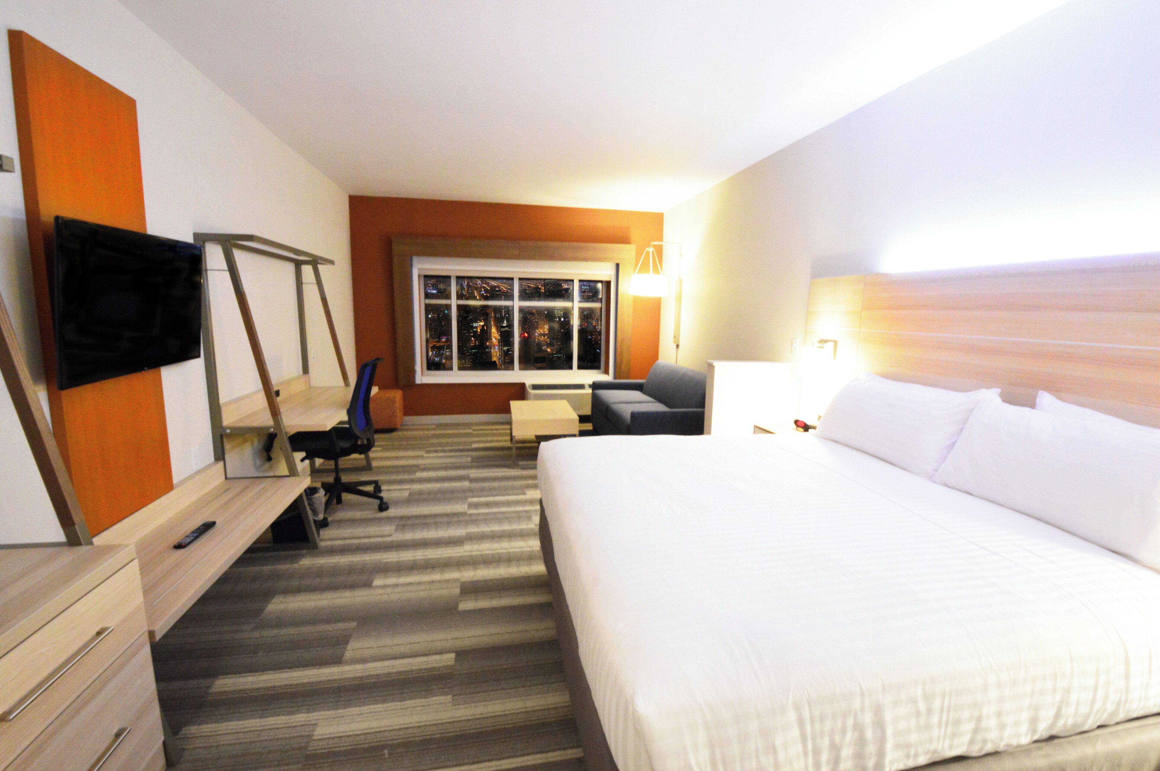 A hotel room with a bed, a desk, a chair, a TV, a window with a city view, and a lamp.