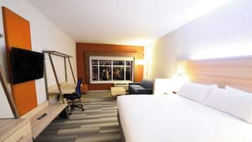 A hotel room with a bed, a desk, a chair, a TV, a window with a city view, and a lamp.