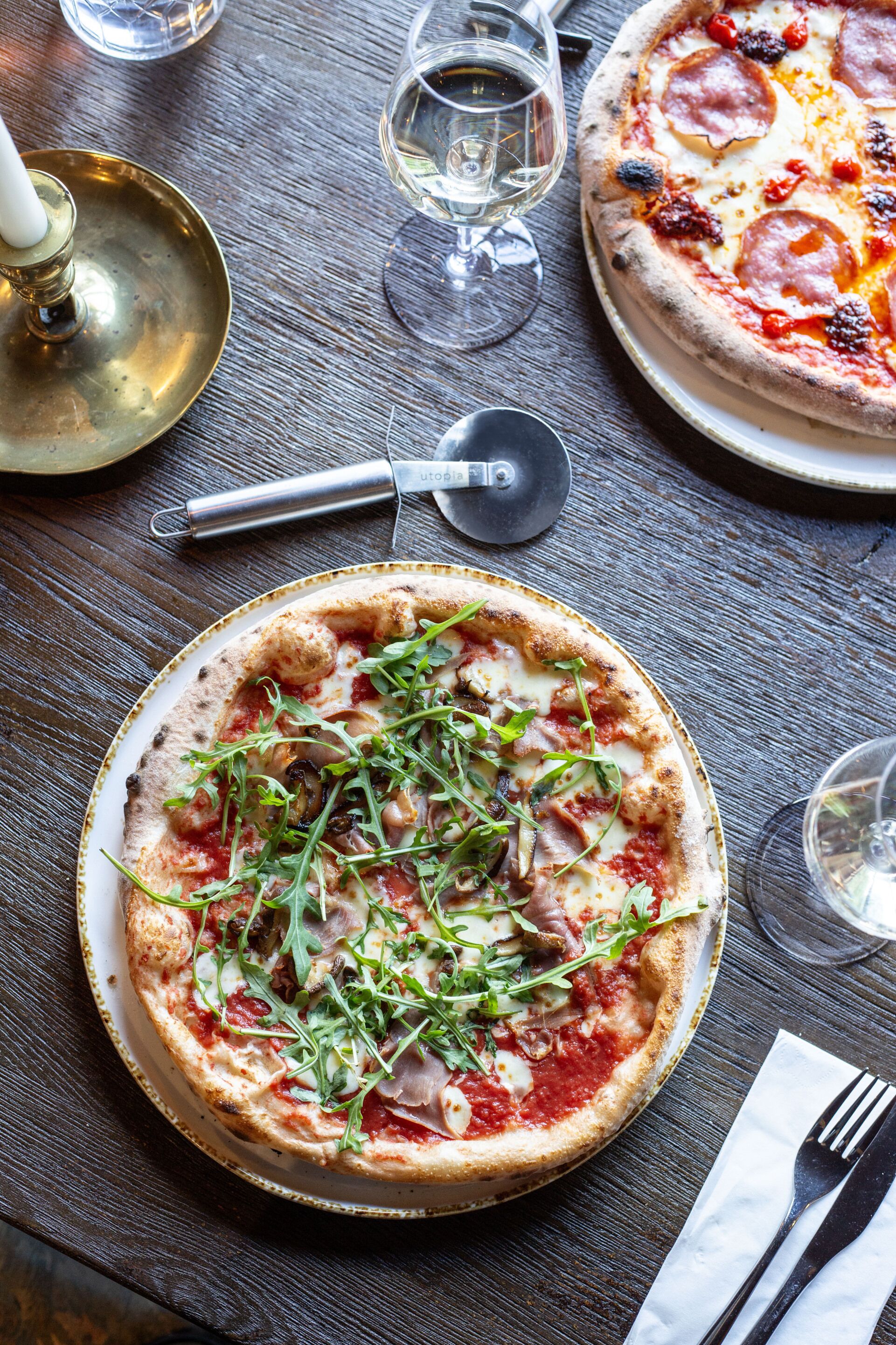 Une pizza garnie de légumes verts et de fromage, présentée sur une assiette, un coupe-pizza et un verre à vin, le tout posé sur une table en bois.