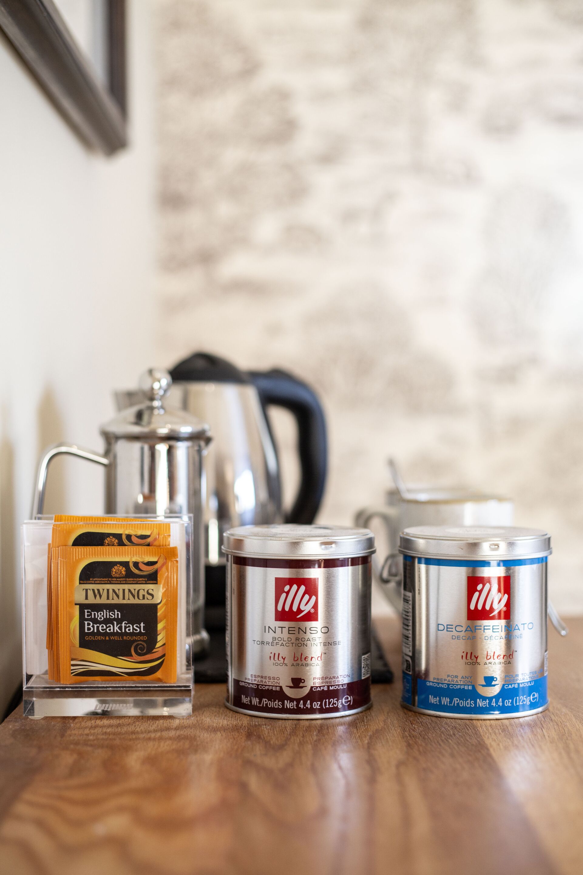 Una cafetera, dos recipientes para café y un paquete de café Twinings English Breakfast sobre una superficie de madera.