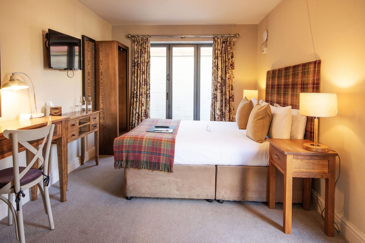 Quarto de hotel com cama, escrivaninha, cadeira, televisão e janela com cortinas.