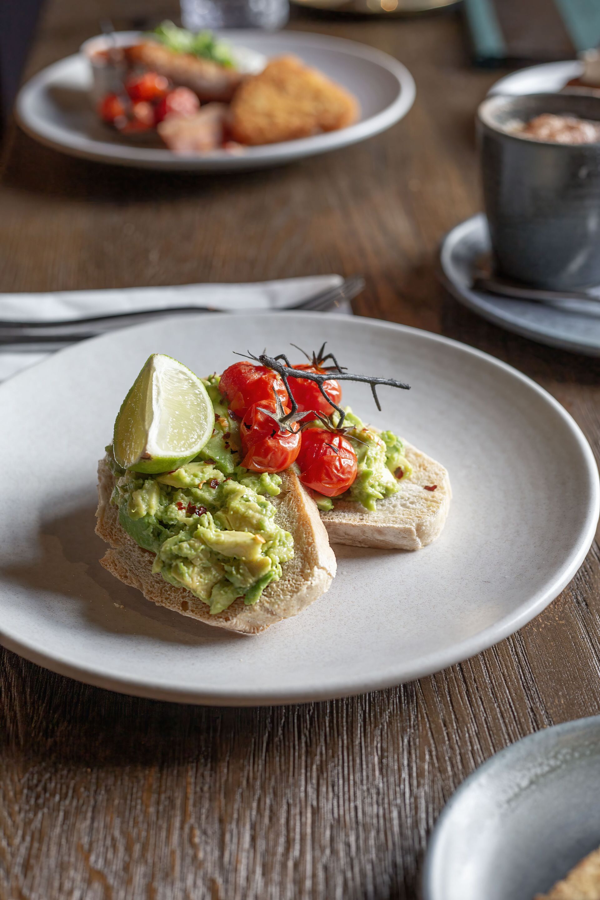 Un plato con tostada de aguacate, tomates cherry y un gajo de lima.