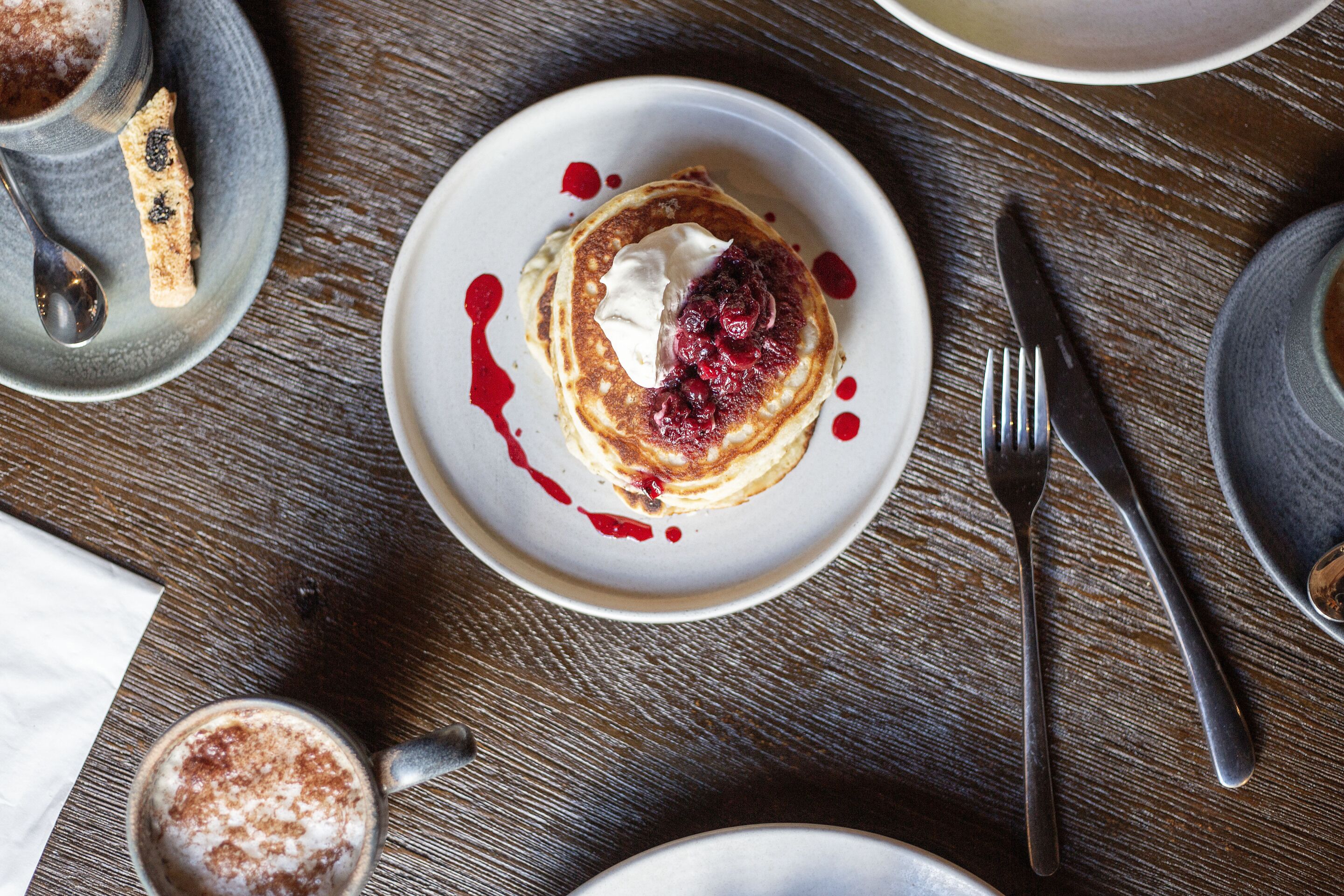 A plate of pancakes with berries and whipped cream, a fork, and a knife on a wooden table.