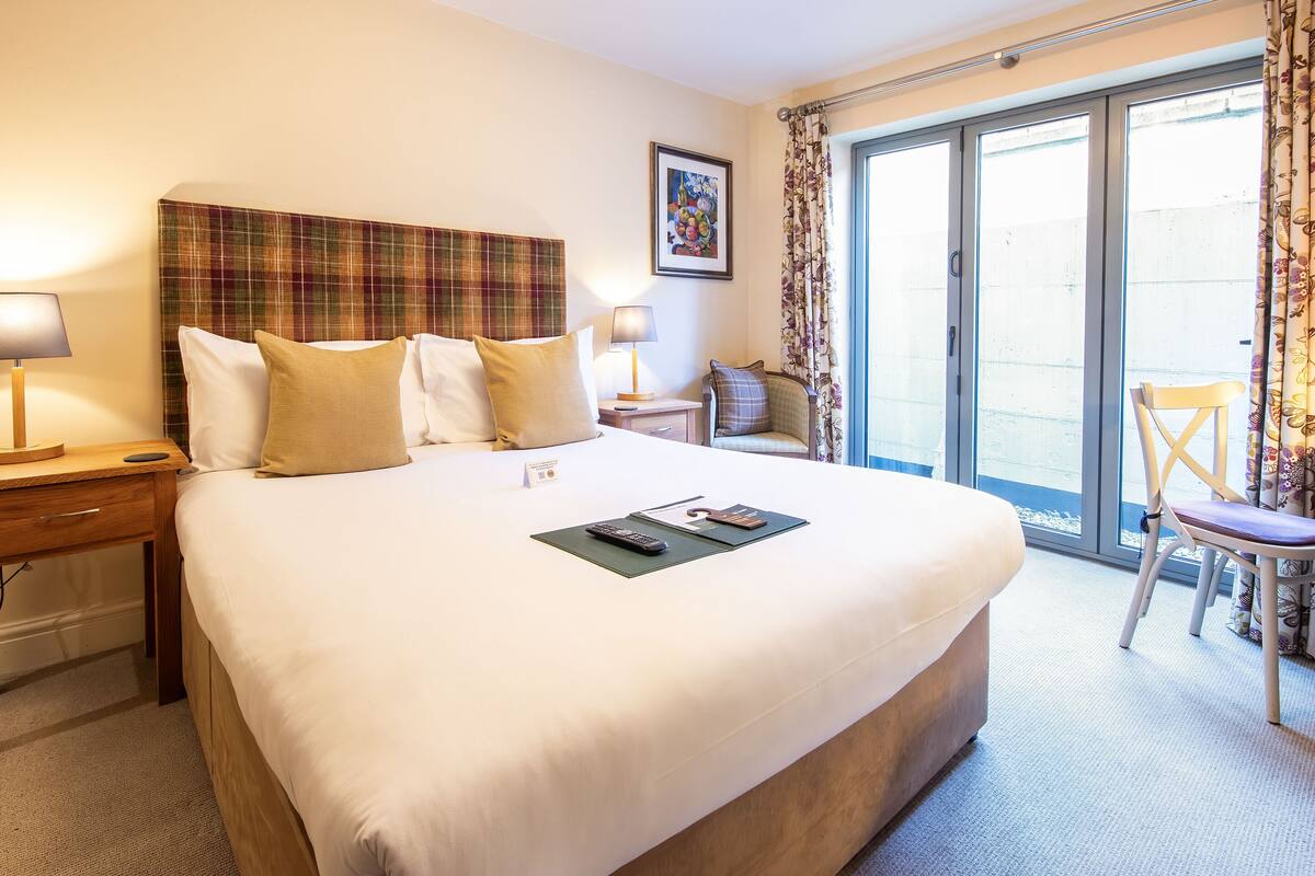 Quarto de hotel com uma cama grande, uma mesa de cabeceira de madeira com abajur, uma cadeira e vista através de uma porta de vidro deslizante.