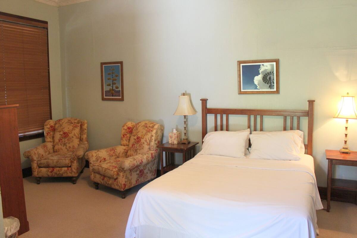 Quarto com cama, duas cadeiras, mesa de cabeceira e duas luminárias de parede.