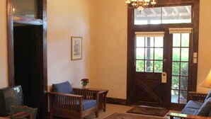 Um quarto com uma mesa de jantar de madeira, duas cadeiras de madeira, um sofá, uma pequena mesa com abajur, um candelabro, uma porta com painéis de vidro e um quadro emoldurado na parede.