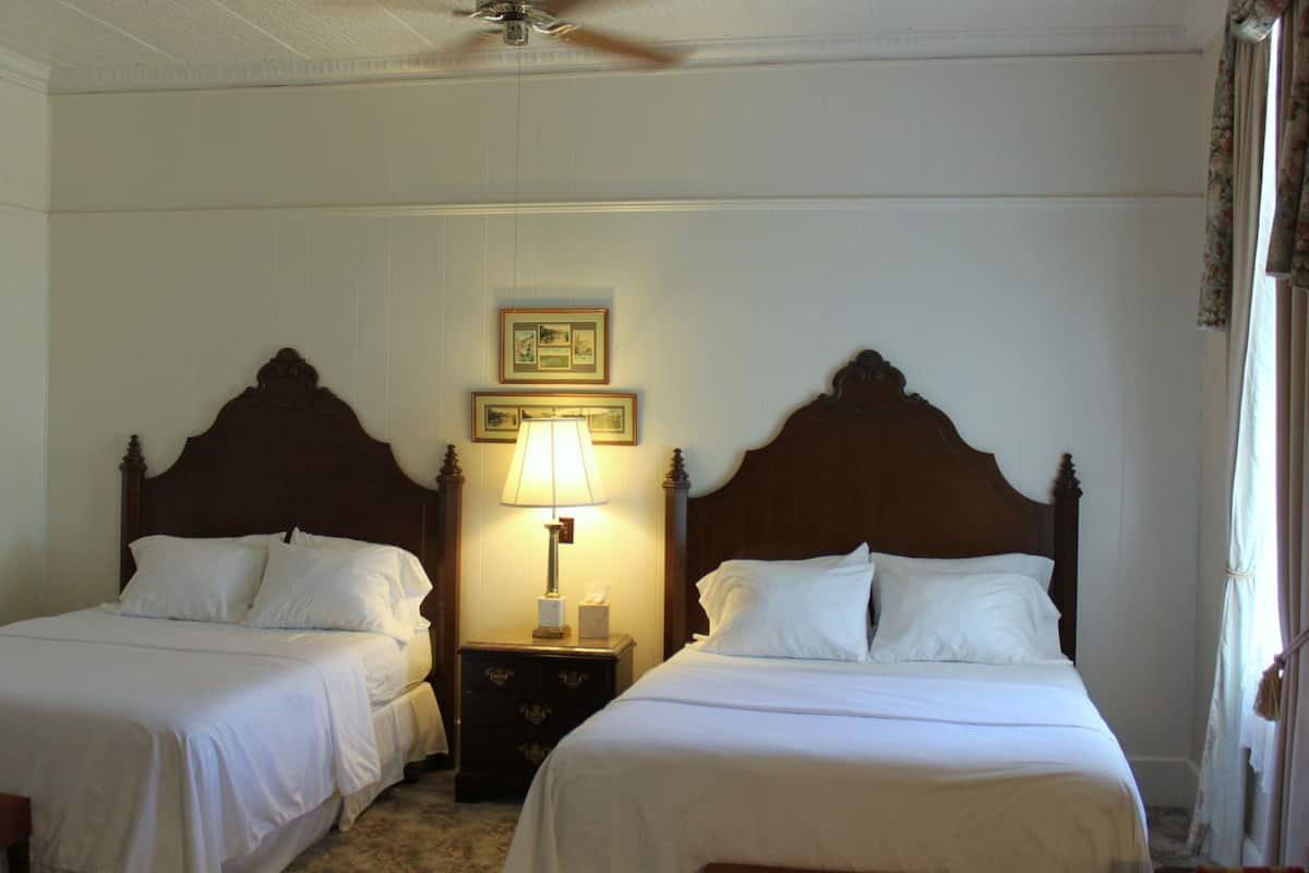 Quarto de hotel com duas camas de solteiro, uma mesa de cabeceira com abajur e um ventilador de teto.