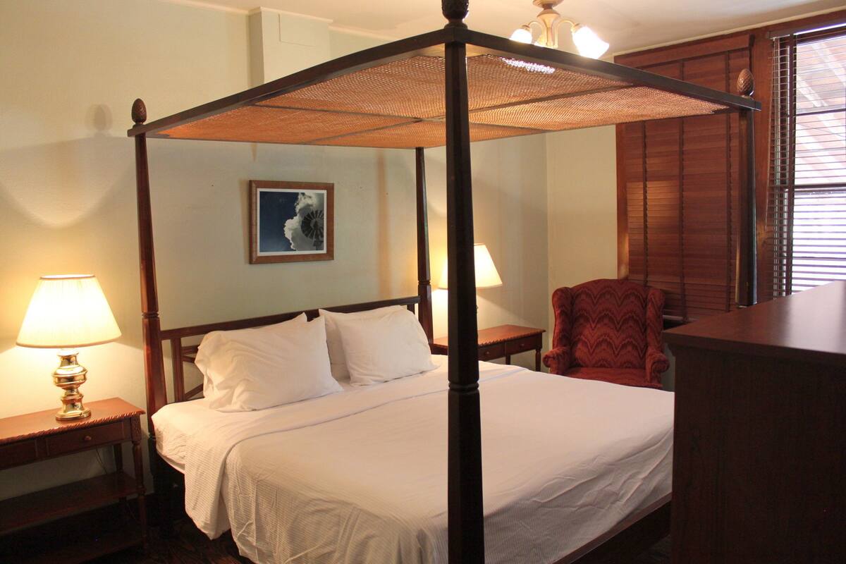 Uma cama de dossel com lençóis brancos, duas mesas de cabeceira com abajures, um quadro na parede e uma cadeira vermelha.