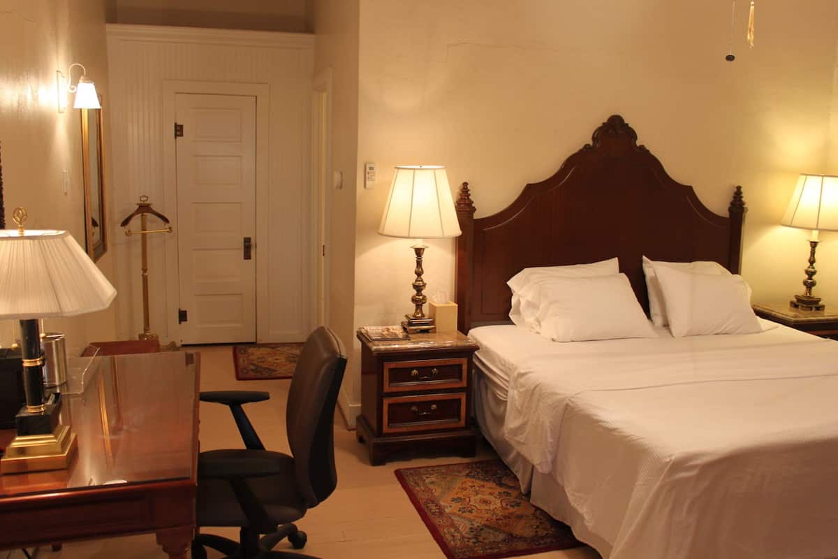 Quarto de hotel com cama de madeira, duas mesas de cabeceira com luminárias, uma escrivaninha com luminária, uma cadeira e uma porta.