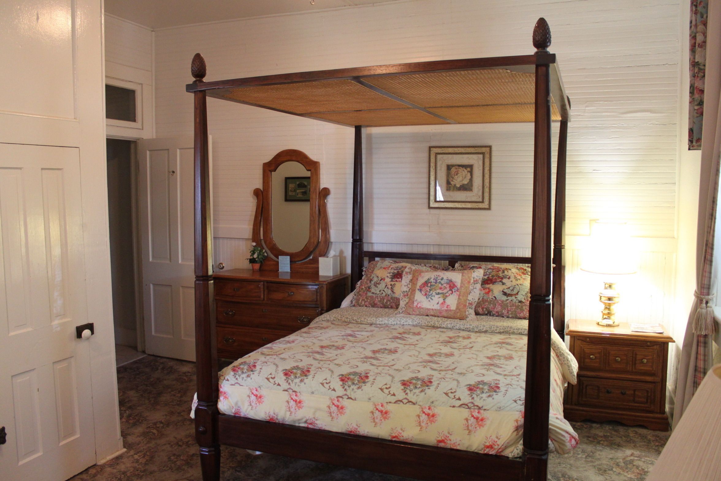 Un lit à baldaquin avec une literie à motifs floraux, une commode en bois avec un miroir et une table de chevet avec une lampe.