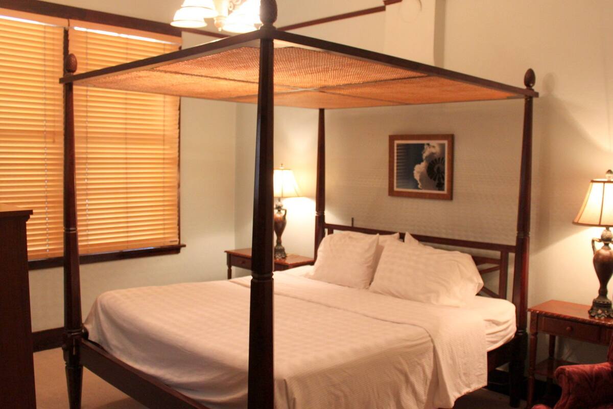 Uma cama com dossel com lençóis brancos, um criado-mudo de madeira com uma lâmpada, um quadro na parede e uma cadeira vermelha.
