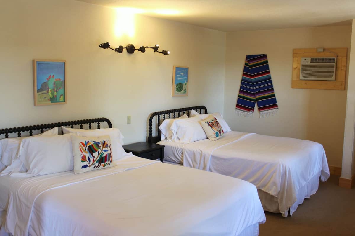 Quarto com duas camas, cada uma com roupa de cama branca e almofadas decorativas. Há um ar condicionado fixado na parede e duas pinturas emolduradas.
