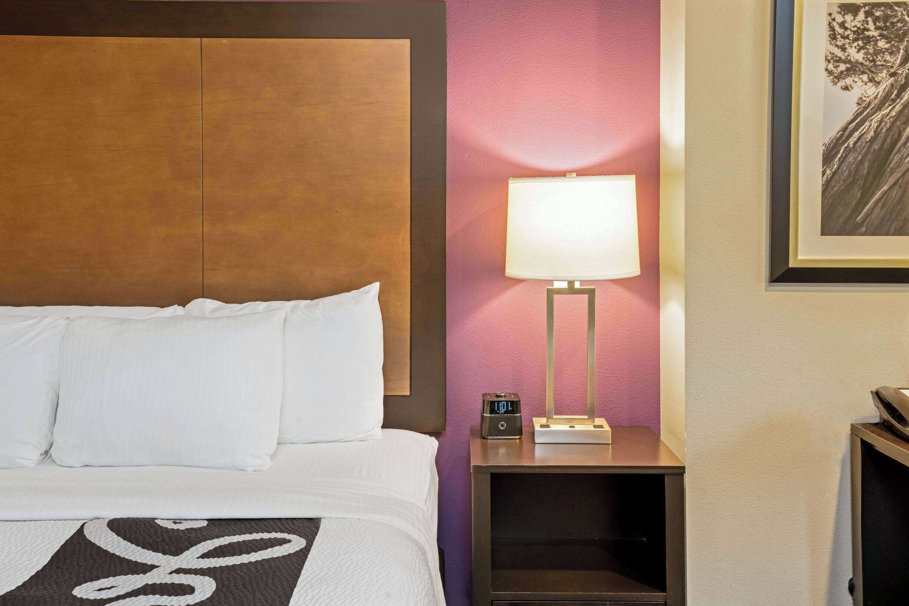 Ein Hotelzimmer mit Bett, Nachttisch mit Lampe, Uhr und einem gerahmten Bild an der Wand.