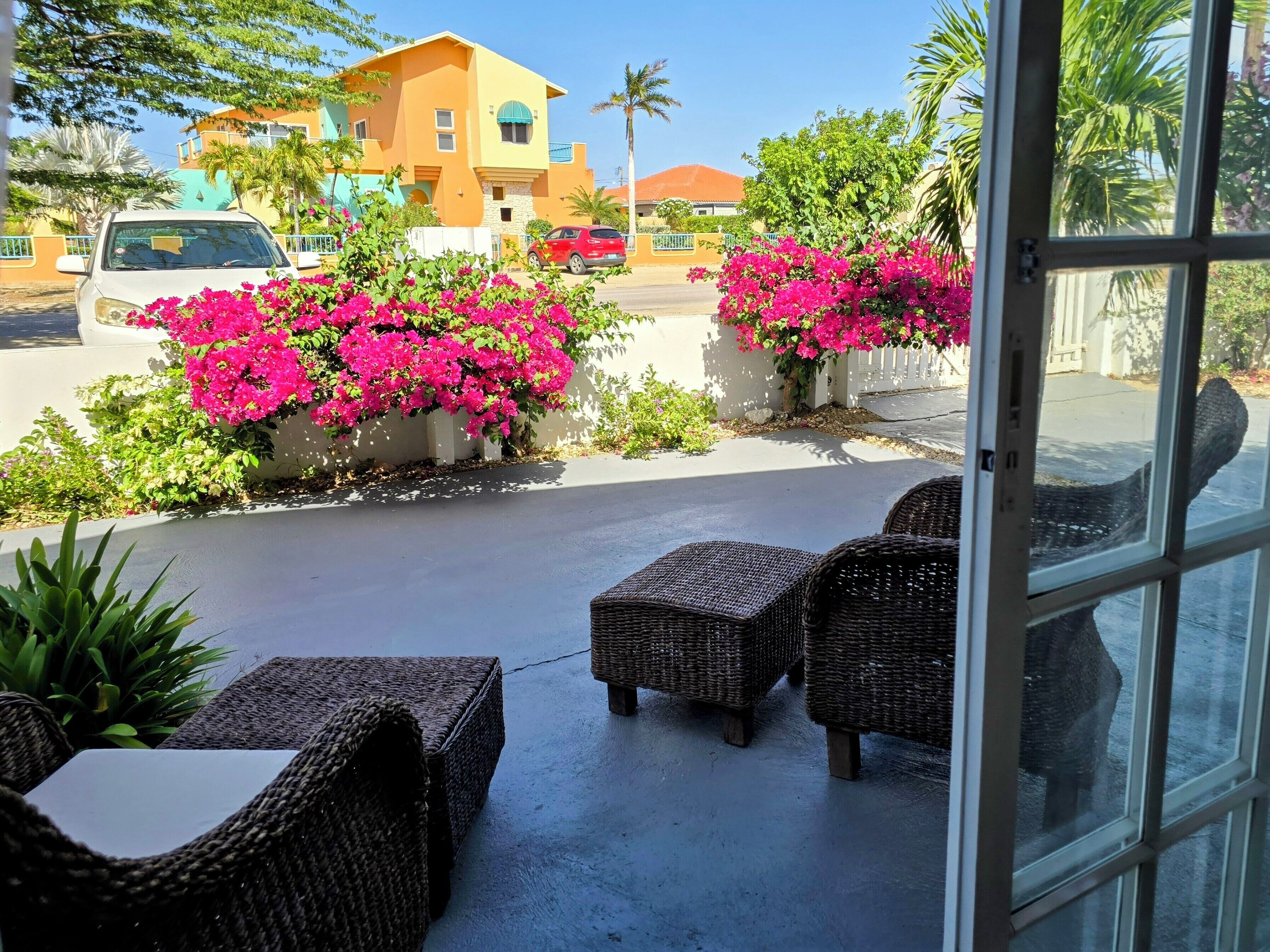 Eine Terrasse mit Korbmöbeln, ein in der Nähe geparktes weißes Auto und leuchtend rosa Bougainvilleen.