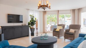 A modern living room with a flat-screen TV, a round coffee table, blue sofas, wicker chairs, and a large window with curtains. - Bubali Villa & Apartments (Oranjestad)