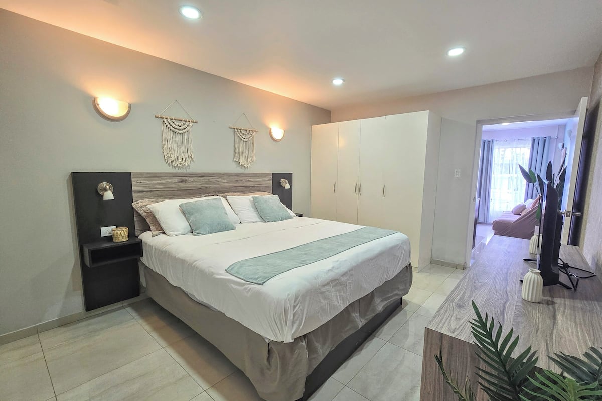 Un dormitorio moderno con una cama grande, mesitas de noche, un armario y un baño visible a través de una puerta abierta.