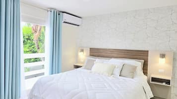 Un dormitorio con una cama grande, cabecera de madera, dos mesitas de noche con lámparas, y vista a la vegetación a través de una ventana con cortinas azules.