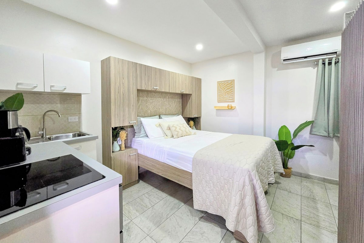 Una habitación de hotel moderna con kitchenette, una cama con cubrecama acolchado, una mesita de noche con una planta y un aire acondicionado de pared.