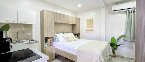 Una habitación de hotel moderna con kitchenette, una cama con cubrecama acolchado, una mesita de noche con una planta y un aire acondicionado de pared.