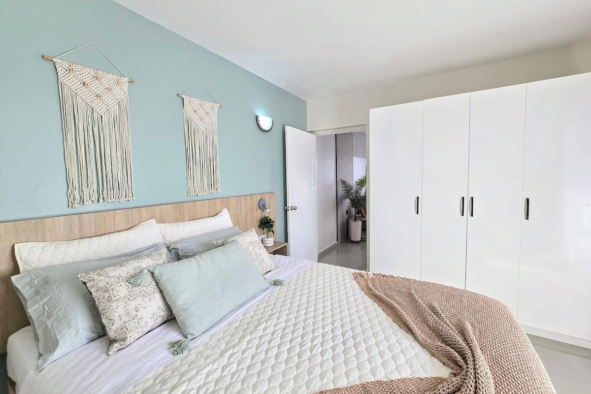 Un dormitorio con cama, almohadas, un colgador de macramé, una lámpara de noche y un armario.