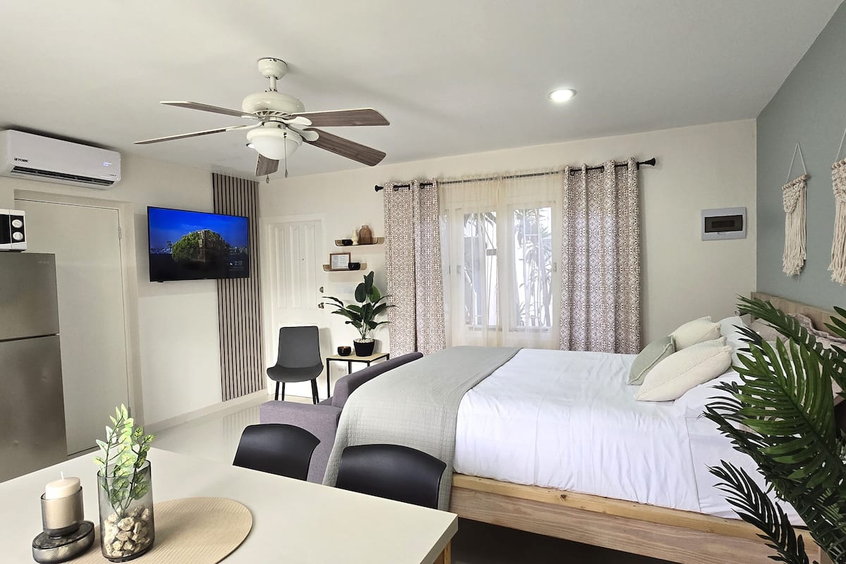 Un dormitorio con cama, mesita de noche, sillas, escritorio, televisor, ventilador de techo y ventana con cortinas.