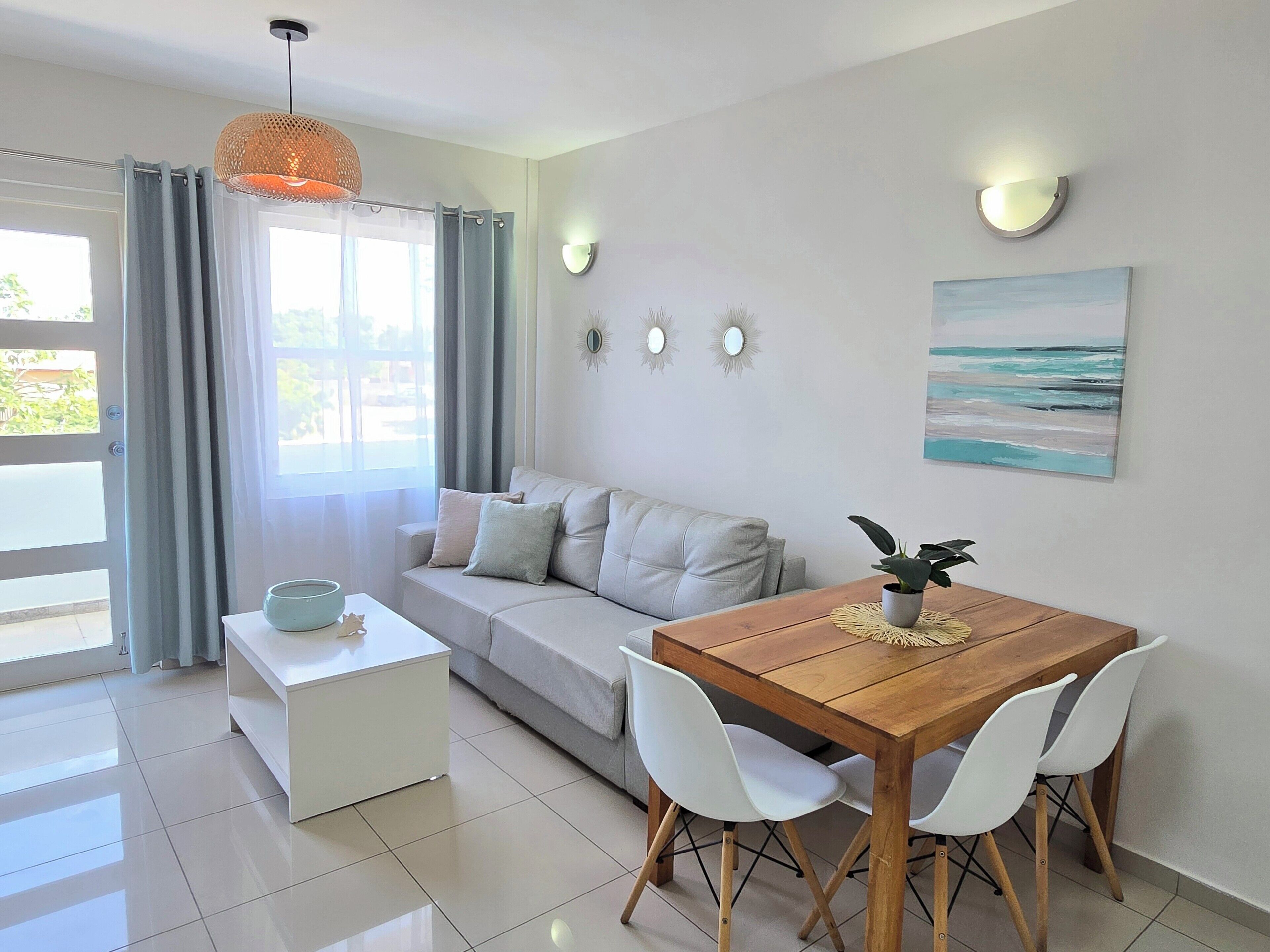 Uma sala de estar moderna com um sofá, uma mesa de jantar de madeira com cadeiras, uma pequena mesa lateral branca e uma grande janela com cortinas.