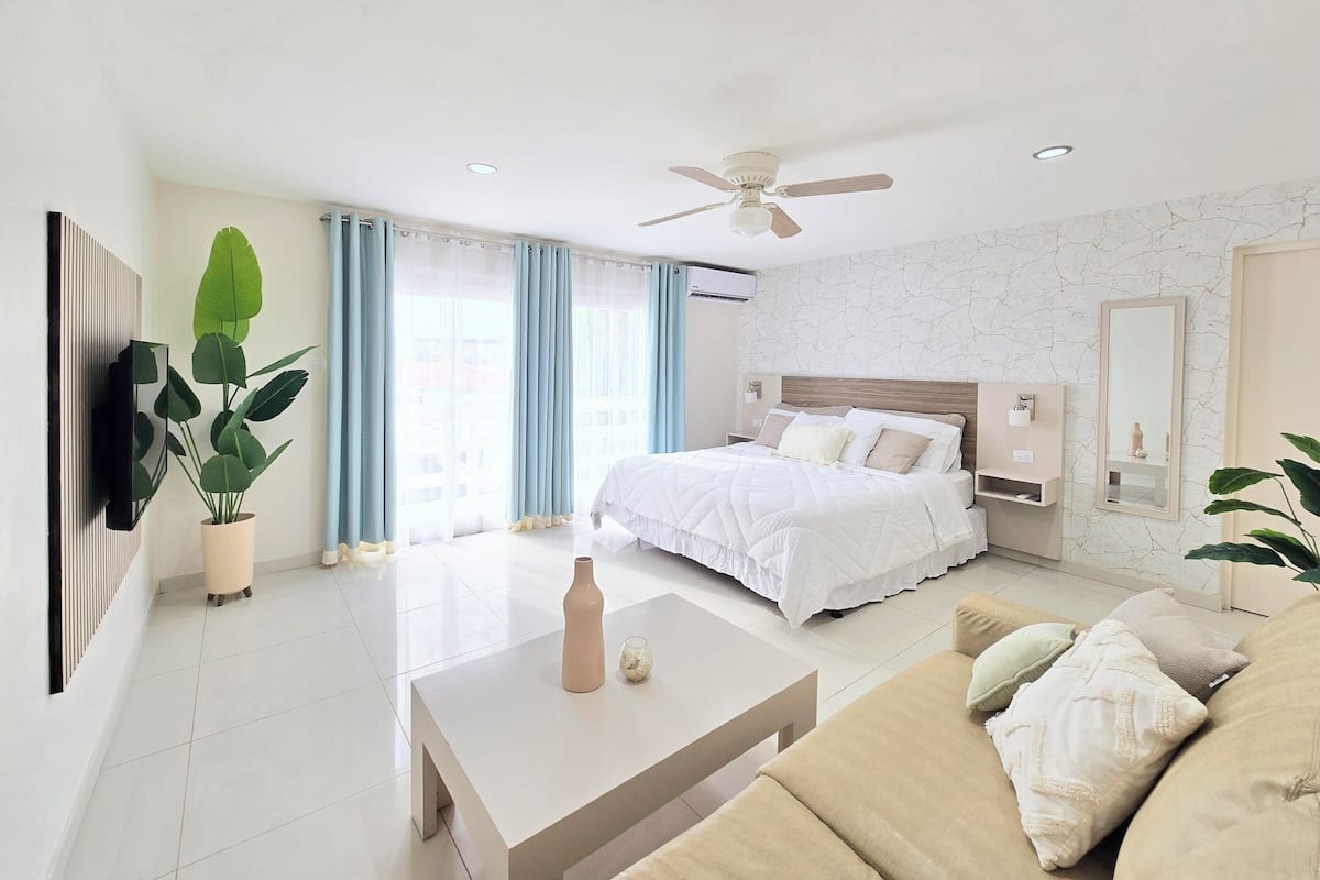Una habitación de hotel moderna con cama, ventilador de techo, televisión y un ventanal con cortinas.