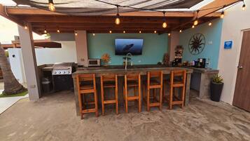 An outdoor kitchen area with a bar, wooden stools, a grill, and a TV screen.