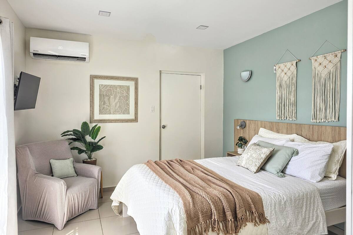 Un dormitorio con cama, una silla, aire acondicionado de pared, un cuadro enmarco y adornos de macramé en la pared.